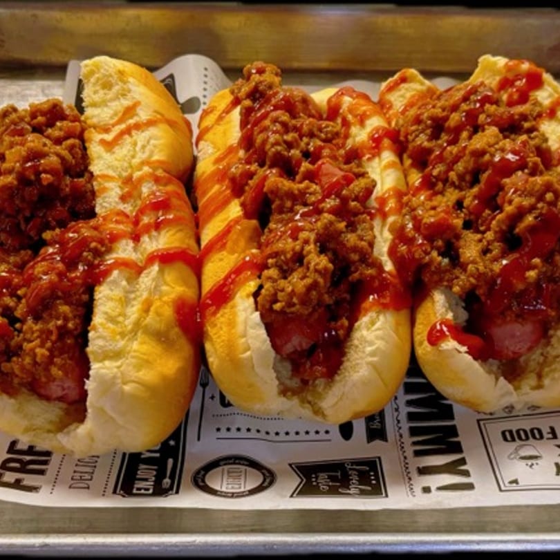 Three chili dogs with ketchup drizzle on a branded paper liner, served on a metal tray.