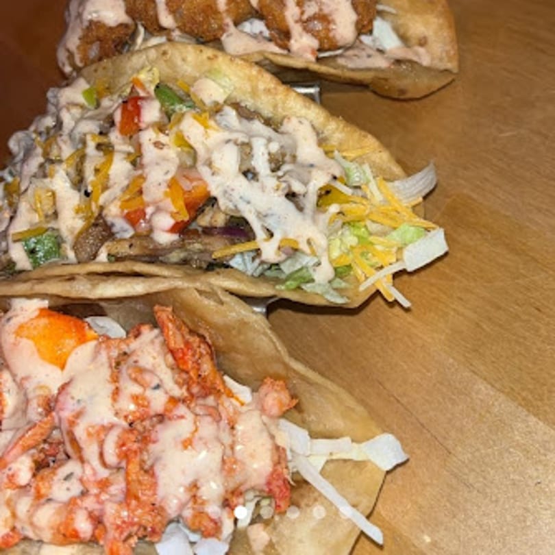 Three loaded tacos with various fillings and sauces, including fried chicken, on a wooden surface.